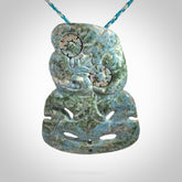 This photo shows a hand carved Aotea stone Tiki pendant. It is a very light blue colour with pāua shell insert eyes. The cord is blue and silver grey and is length adjustable